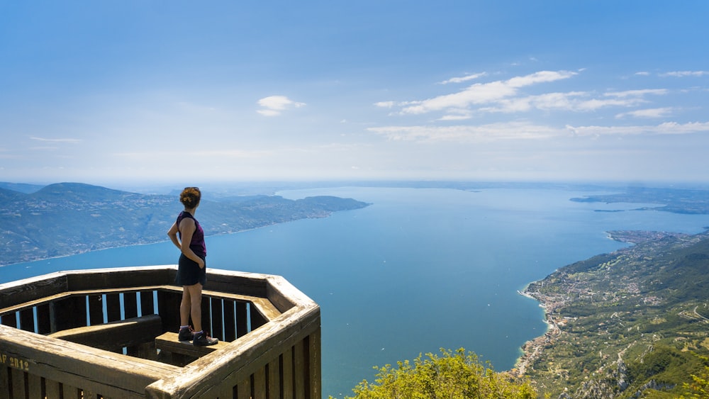 Frau im schwarzen Tanktop steht in der Nähe des Geländers und sieht tagsüber blaues Meer und Berge unter blauem und weißem Himmel