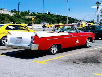 A vintage red and white convertible car is parked in a parking lot with a scenic backdrop of green hills and a partly cloudy sky. Several other cars, including a yellow taxi, are visible nearby, and there are lamp posts and palm trees lining the street.