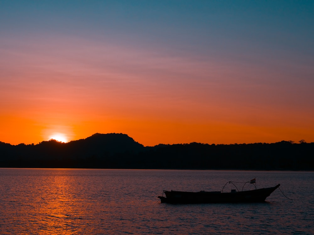 silhouette of boat on calm water by mountain during golden hour