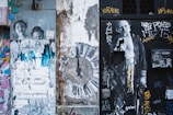The image features a distressed urban wall covered in various street art elements including graffiti, stenciled images, and posters. On the left, there are partially torn posters of people, and in the center, a large clock design is painted on the wall. On the right, a black door is covered with more graffiti and another image of a person. The overall feel is grungy with layers of art and decay.