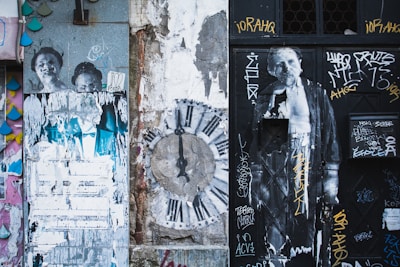 The image features a distressed urban wall covered in various street art elements including graffiti, stenciled images, and posters. On the left, there are partially torn posters of people, and in the center, a large clock design is painted on the wall. On the right, a black door is covered with more graffiti and another image of a person. The overall feel is grungy with layers of art and decay.