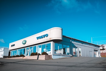 A car dealership building with clear signage for Volvo, Jaguar, and Land Rover brands. The structure features large glass windows and a metallic exterior, situated on a slightly elevated area with a clear, blue sky in the background.