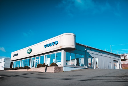 A car dealership building with clear signage for Volvo, Jaguar, and Land Rover brands. The structure features large glass windows and a metallic exterior, situated on a slightly elevated area with a clear, blue sky in the background.