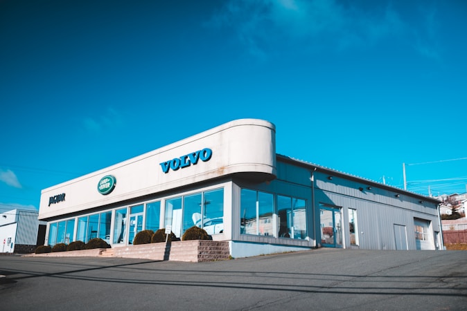 A car dealership building with clear signage for Volvo, Jaguar, and Land Rover brands. The structure features large glass windows and a metallic exterior, situated on a slightly elevated area with a clear, blue sky in the background.
