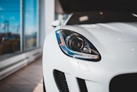 A close-up of the front left headlight of a sleek, modern white car, positioned in a well-lit showroom with large windows.