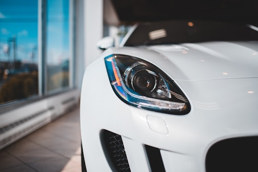 A close-up of the front left headlight of a sleek, modern white car, positioned in a well-lit showroom with large windows.