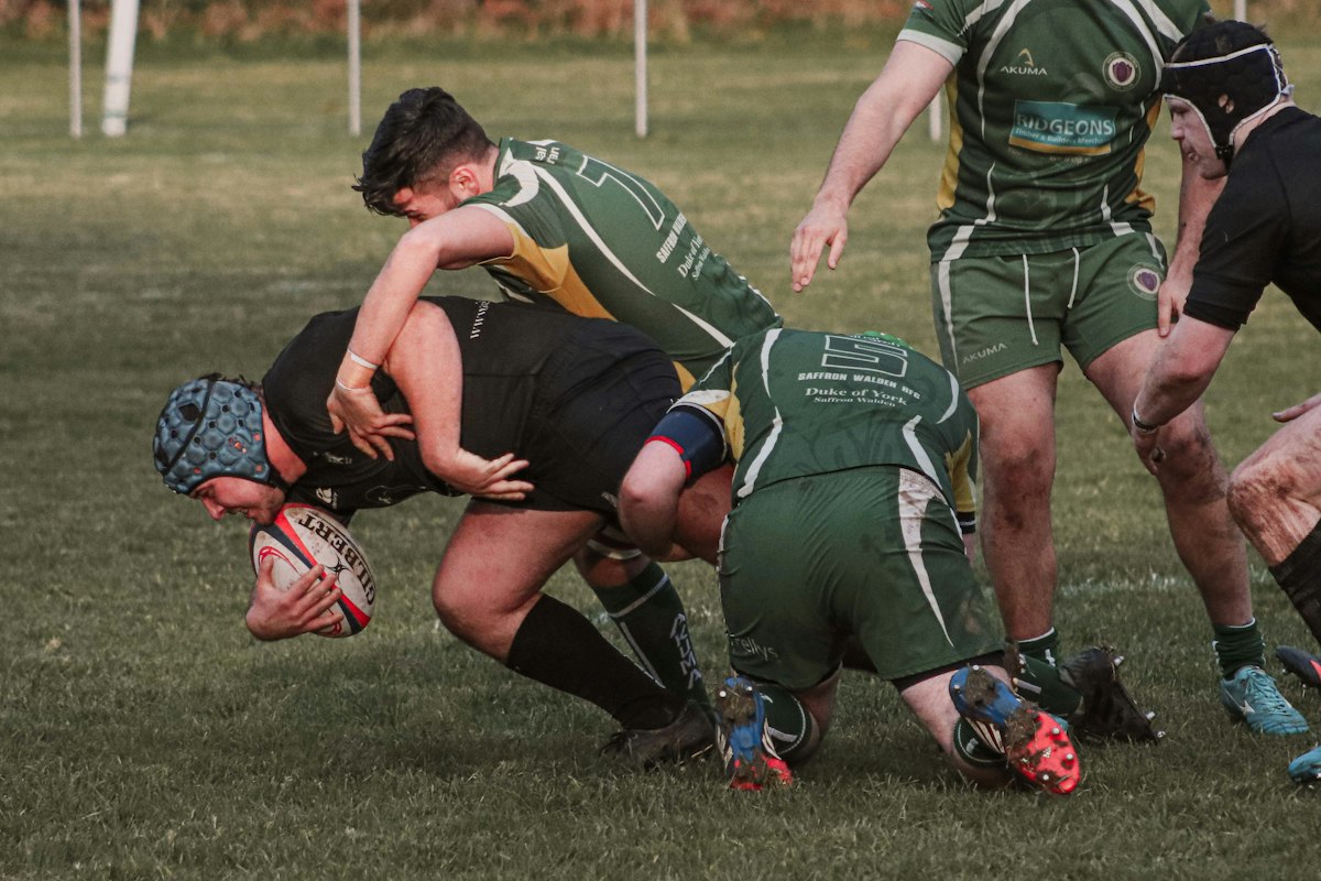 Plaquage au ras du sol dans un match de rugby amateur, un joueur en maillot vert tenant le ballon sous un défenseur en noir