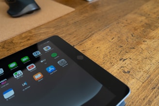 A tablet displaying various app icons is placed on a wooden table. Nearby, a partially visible computer mouse rests on a mouse pad.