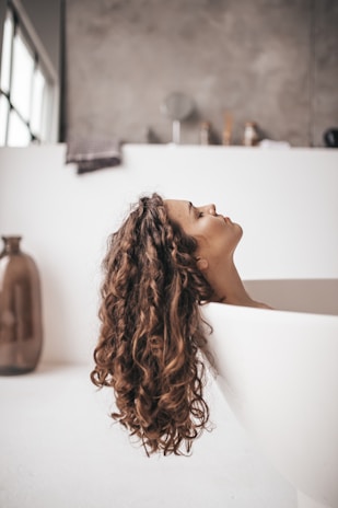 A peaceful moment of self-care: a woman smiling while rinsing her hair in a sunlit bathroom.