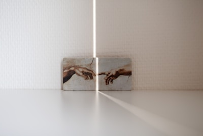 A small artwork is displayed against a white textured wall, featuring the iconic image of two hands reaching out to each other. The lighting creates a soft shadow and ray of light pointing towards the artwork, adding an ethereal quality to the scene.