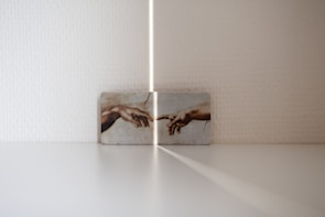 A small artwork is displayed against a white textured wall, featuring the iconic image of two hands reaching out to each other. The lighting creates a soft shadow and ray of light pointing towards the artwork, adding an ethereal quality to the scene.