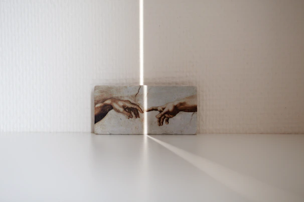 A small artwork is displayed against a white textured wall, featuring the iconic image of two hands reaching out to each other. The lighting creates a soft shadow and ray of light pointing towards the artwork, adding an ethereal quality to the scene.