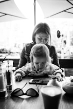 A black and white image depicting a young child intently looking at a smartphone on a wooden table, with an adult, possibly a parent, seated behind them. The setting appears to be a cafe or restaurant with blurred outdoor elements in the background. The table also holds a pair of glasses, salt and pepper shakers, and a coffee cup.