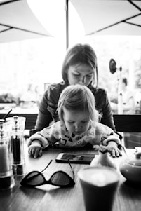 A black and white image depicting a young child intently looking at a smartphone on a wooden table, with an adult, possibly a parent, seated behind them. The setting appears to be a cafe or restaurant with blurred outdoor elements in the background. The table also holds a pair of glasses, salt and pepper shakers, and a coffee cup.