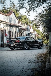 A black sports car is parked on a cobblestone street lined with charming, white Spanish-style houses. The houses have curved tile roofs and arched windows. The street is partially shaded by overhanging branches and surrounded by lush green foliage.