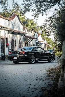 A black sports car is parked on a cobblestone street lined with charming, white Spanish-style houses. The houses have curved tile roofs and arched windows. The street is partially shaded by overhanging branches and surrounded by lush green foliage.