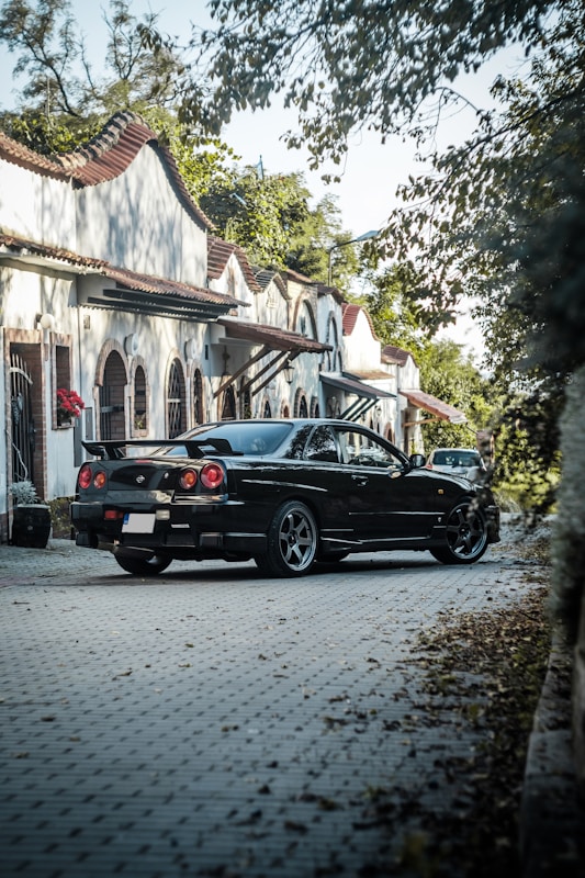 A black sports car is parked on a cobblestone street lined with charming, white Spanish-style houses. The houses have curved tile roofs and arched windows. The street is partially shaded by overhanging branches and surrounded by lush green foliage.