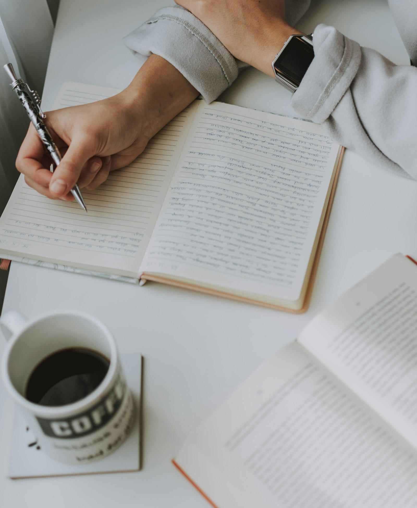 Scrivania con quaderno aperto, mano che scrive e una tazza di caffè