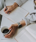 A person writing content on a notepad with a coffee cup.