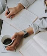 A person is sitting at a desk, writing in a lined notebook with a pen in one hand. The other hand is holding a cup of coffee, while an open book lies on the desk nearby. The individual is wearing a watch and a light-colored garment.