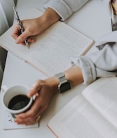 A person is sitting at a desk, writing in a lined notebook with a pen in one hand. The other hand is holding a cup of coffee, while an open book lies on the desk nearby. The individual is wearing a watch and a light-colored garment.