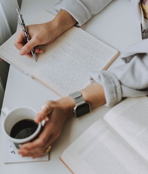 A person is sitting at a desk, writing in a lined notebook with a pen in one hand. The other hand is holding a cup of coffee, while an open book lies on the desk nearby. The individual is wearing a watch and a light-colored garment.