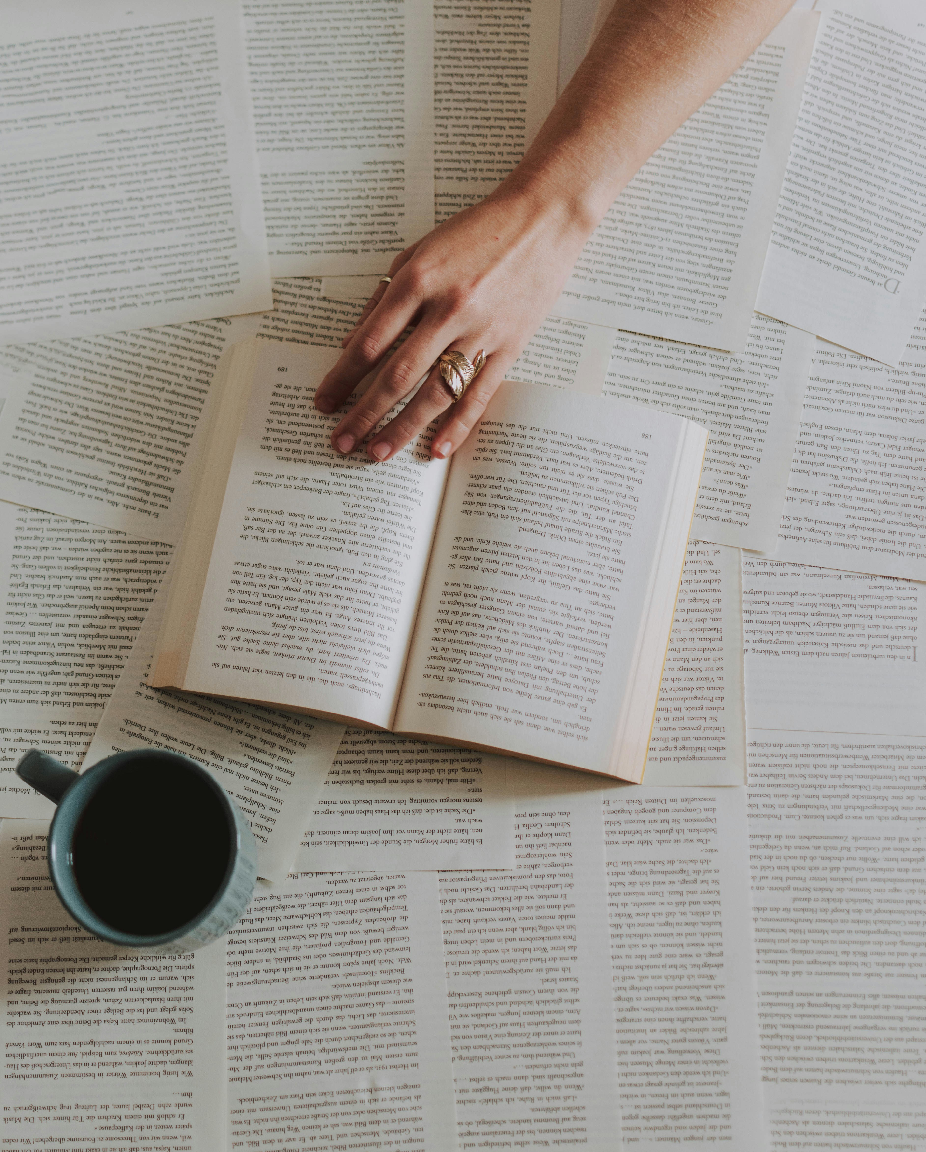 A flat lay of an open book and scattered printed pages on a table, with a mug of dark beverage.