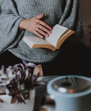 A person wearing a cozy grey sweater is holding an open book, suggesting a relaxed reading moment. In the foreground, there is a soft-focus image of a small potted plant and a light-colored ceramic cup.