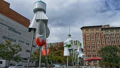 Three abstract metal sculptures with a futuristic design, featuring vibrant red and green accents. The sculptures are situated in an urban environment with distinctive modern and classical buildings in the background. Trees and a sidewalk caf&eacute; with red umbrellas add context to the city setting under a cloudy sky.