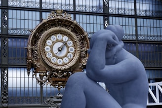 A large ornate clock with Roman numerals sits against a backdrop of a grid-like window. In the foreground, a blue-toned sculpture of a person is posed with their head resting on their hand.