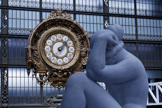 A large ornate clock with Roman numerals sits against a backdrop of a grid-like window. In the foreground, a blue-toned sculpture of a person is posed with their head resting on their hand.