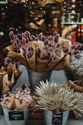 A display of various bouquets in metal buckets, featuring a range of dried flowers, including purple thistles, light pink blossoms, and beige palm leaves. Brown paper wraps several of the bouquets, and chalkboard signs indicate prices. The background is softly blurred, with hints of twinkling lights, suggesting a cozy, inviting atmosphere.