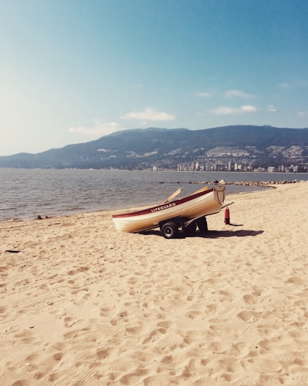 A lifeguard boat is stationed on a sandy beach, slightly tilted on a trailer. The boat has the word 'LIFEGUARD' written on its side. A fire extinguisher is placed near the boat. In the background, there is a large body of water, possibly a lake or sea, with mountains and a cityscape visible in the distance under a clear blue sky.