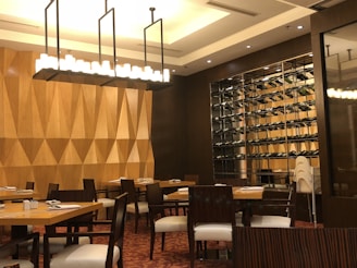 A restaurant interior with wooden geometric patterned wall panels and a large rectangular lighting fixture hanging from the ceiling. There are several tables and chairs arranged neatly. A wall-mounted wine rack filled with wine bottles is prominently displayed. The floor is carpeted in a patterned red design, and the general ambiance is warm and inviting.