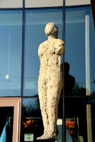 A textured stone sculpture of a human figure stands against a glass building facade. The surface of the sculpture appears rough and uneven, reflecting light in various spots. The building behind features large window panels, and there is a reflection of the sculpture on the glass. Below, there are clothing items visible through the windows of the building.