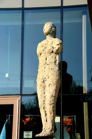 A textured stone sculpture of a human figure stands against a glass building facade. The surface of the sculpture appears rough and uneven, reflecting light in various spots. The building behind features large window panels, and there is a reflection of the sculpture on the glass. Below, there are clothing items visible through the windows of the building.