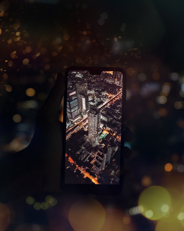 A hand holds a smartphone displaying a vibrant nighttime cityscape with brightly lit skyscrapers and a busy street. The background is blurred with bokeh lights, creating a contrast between the sharpness of the cityscape and the soft, warm-colored circles of light.