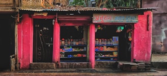 A small street shop with bright pink walls is filled with various goods. The shop has two open sections displaying items like packaged goods, jars, and other grocery store products. The storefront is rugged with some bricks and a paved sidewalk in front.