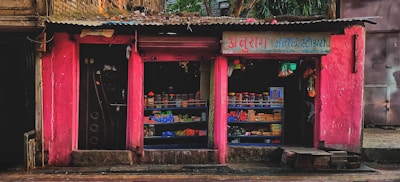 A small street shop with bright pink walls is filled with various goods. The shop has two open sections displaying items like packaged goods, jars, and other grocery store products. The storefront is rugged with some bricks and a paved sidewalk in front.