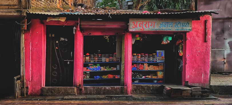 A small street shop with bright pink walls is filled with various goods. The shop has two open sections displaying items like packaged goods, jars, and other grocery store products. The storefront is rugged with some bricks and a paved sidewalk in front.