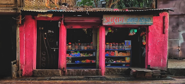 A small street shop with bright pink walls is filled with various goods. The shop has two open sections displaying items like packaged goods, jars, and other grocery store products. The storefront is rugged with some bricks and a paved sidewalk in front.