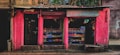 A small street shop with bright pink walls is filled with various goods. The shop has two open sections displaying items like packaged goods, jars, and other grocery store products. The storefront is rugged with some bricks and a paved sidewalk in front.