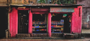 A small street shop with bright pink walls is filled with various goods. The shop has two open sections displaying items like packaged goods, jars, and other grocery store products. The storefront is rugged with some bricks and a paved sidewalk in front.