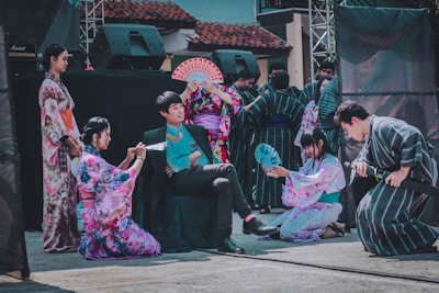 A group of people dressed in traditional Japanese clothing, including kimonos and yukatas, are performing in an outdoor setting. One person is seated while others are kneeling or standing around him with accessories like fans. The backdrop includes a stage with speakers.