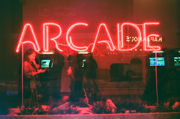 Bright neon red letters spelling out 'ARCADE' dominate the scene. Below the sign, people are reflected in the window, adding a sense of activity and interaction. Inside, arcade machines with glowing screens hint at a bustling entertainment environment.