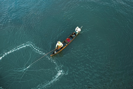 A small wooden boat is being rowed by two individuals on a vast expanse of blue-green water. One person is actively engaged in casting a fishing net, which is spread out in a semicircular pattern on the water’s surface. The scene captures a moment of traditional fishing activity.