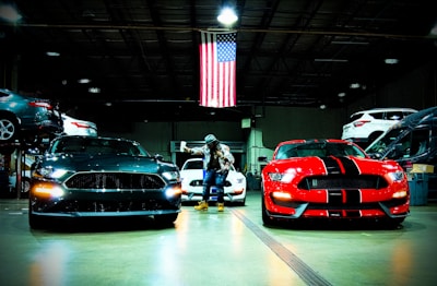 A garage with two prominent sports cars, one black and one red with black stripes, under bright lights. An American flag hangs overhead, and a person stands between the cars. There are other vehicles visible in the background, some elevated on lifts.