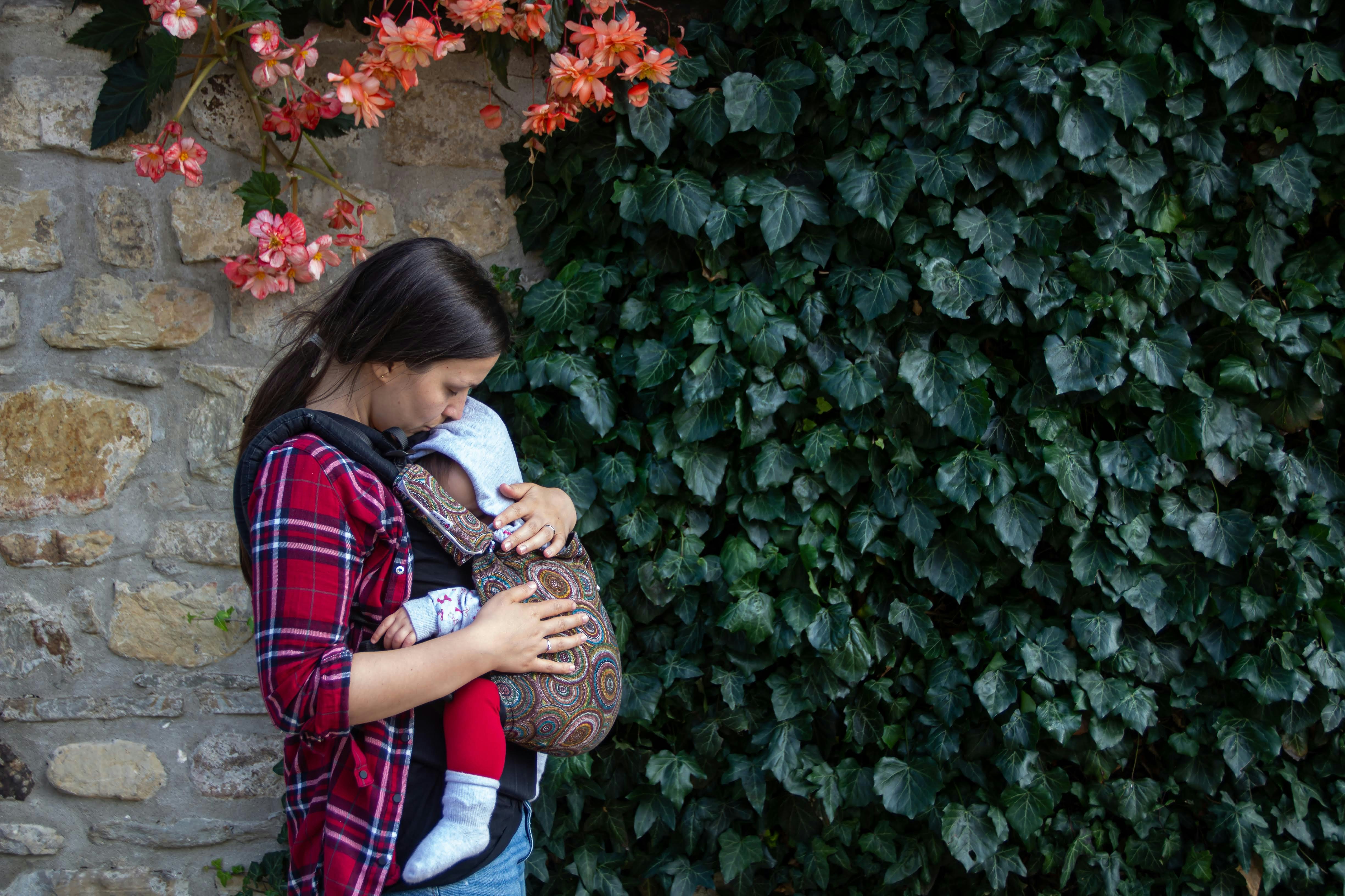 Mother in a plaid shirt carrying her baby facing in, in a structured carrier, beside an ivy-covered stone wall