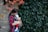 A mother is gently holding a baby in a patterned carrier against a backdrop of lush green ivy and a stone wall. Red and pink flowers add a splash of color to the scene, creating a serene and tender moment.