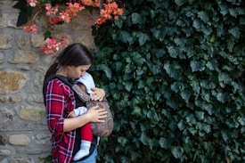 A mother is gently holding a baby in a patterned carrier against a backdrop of lush green ivy and a stone wall. Red and pink flowers add a splash of color to the scene, creating a serene and tender moment.